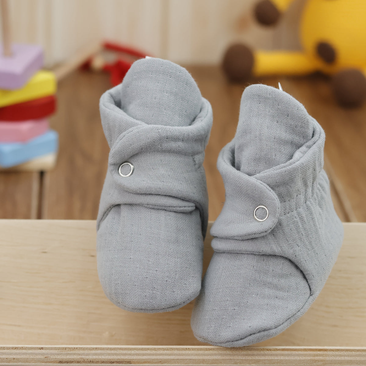 Front view of a pair of light gray textured muslin baby booties with snap straps.