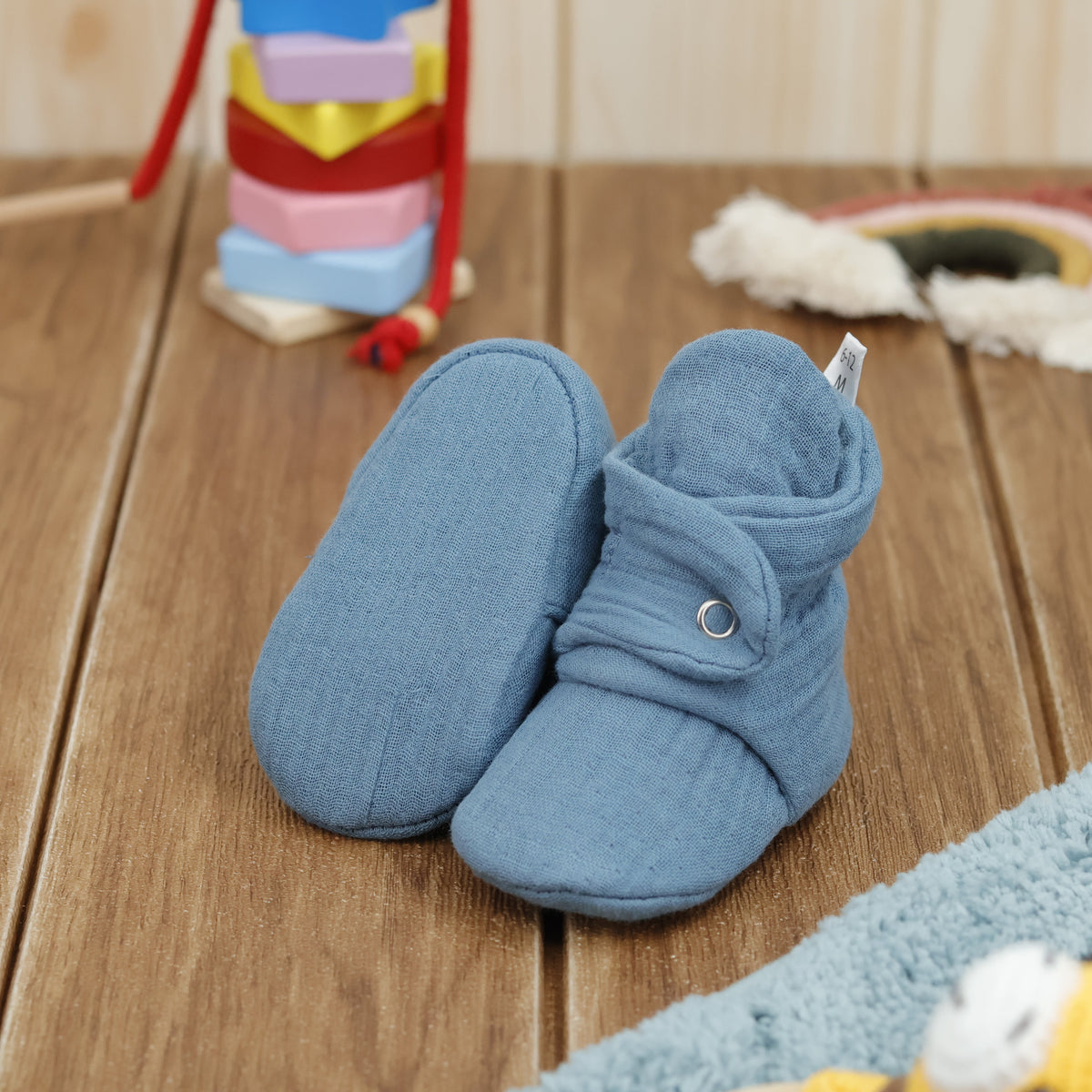 Muted blue textured muslin baby booties with snap straps arranged on a surface with one sole facing the camera.