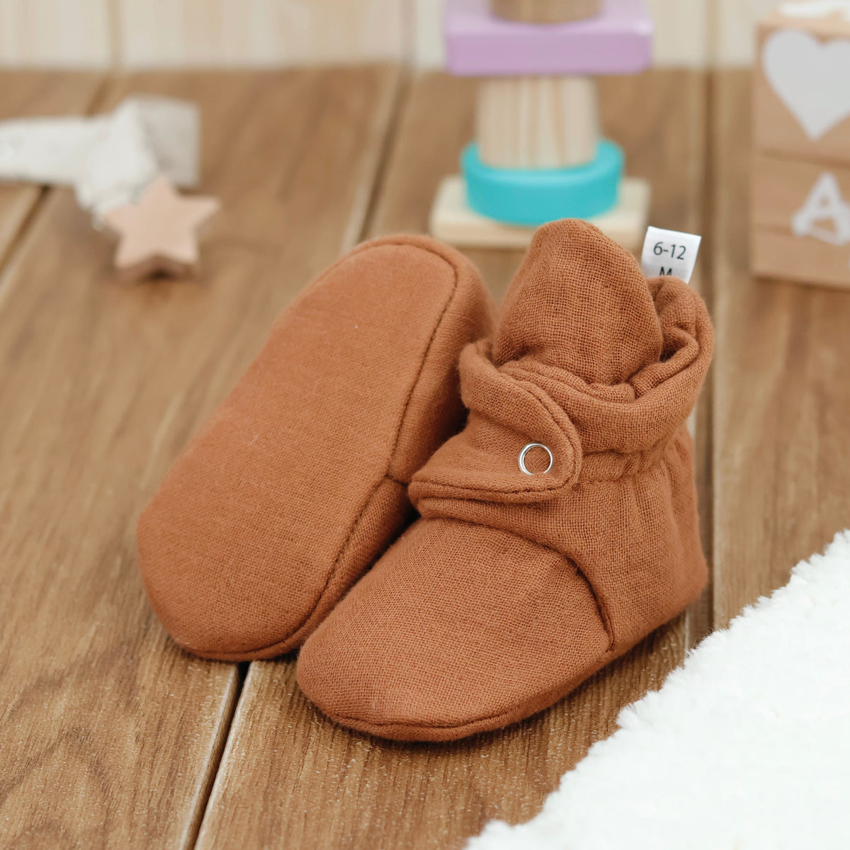Warm brown textured muslin baby booties with snap straps arranged on a surface with one sole facing the camera.