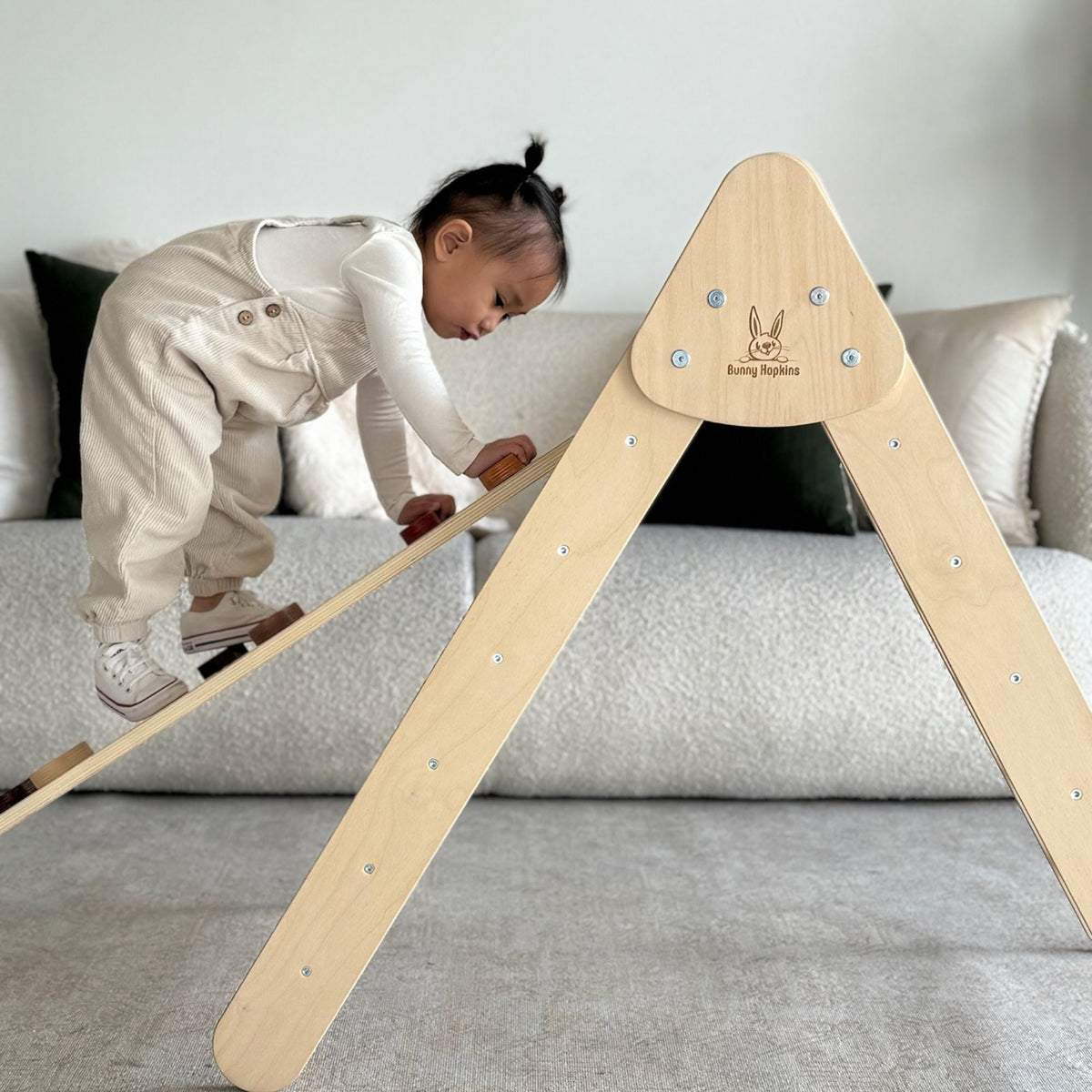 Child climbing a ramp attached to a pale natural-tone triangle beside a sofa.