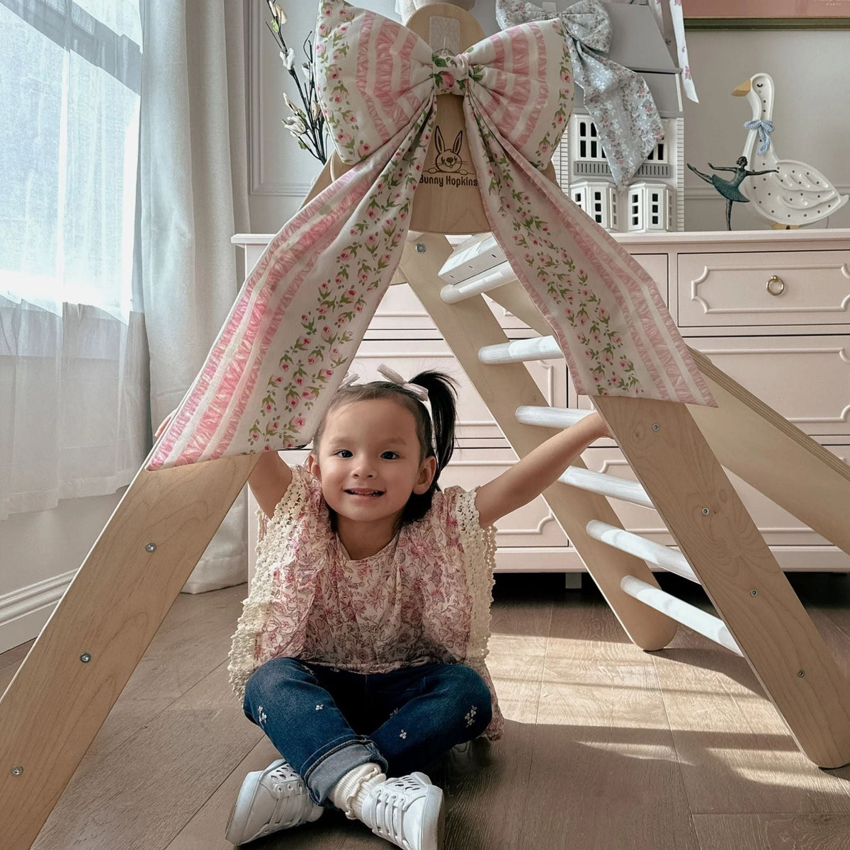 Child seated beneath a white-rung climbing triangle decorated with a floral fabric bow.
