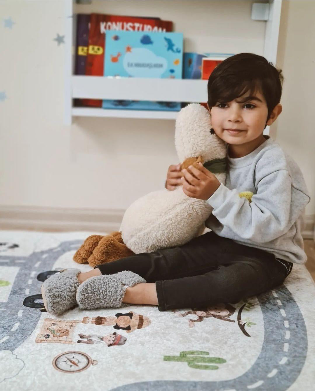 Child seated on a rug holding a teddy bear and wearing gray teddy booties.
