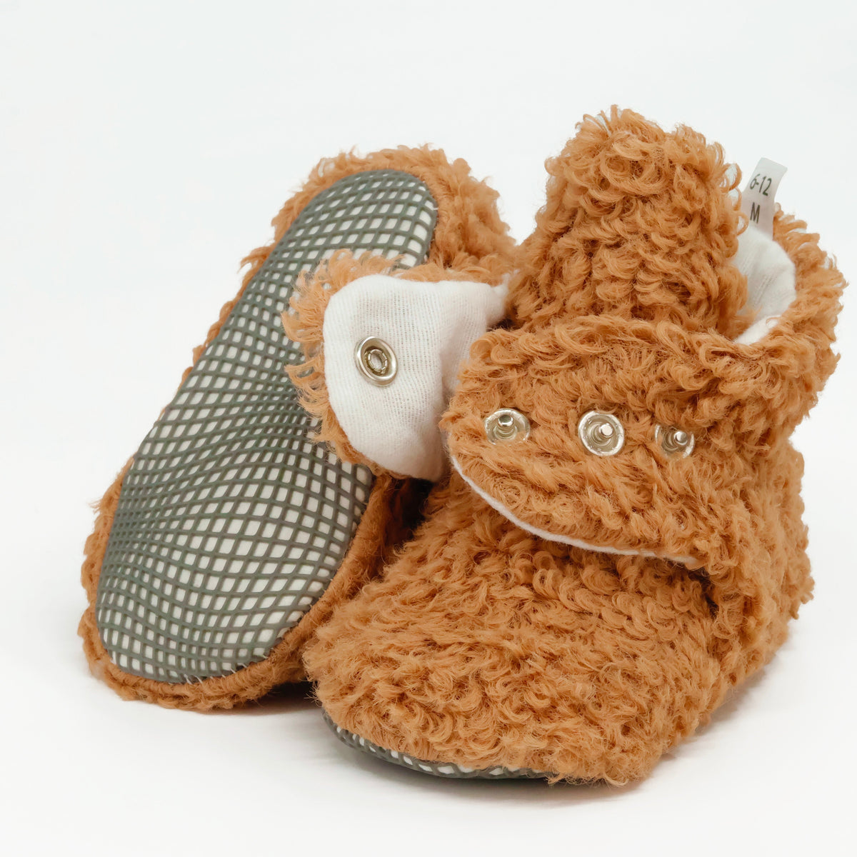 Light brown teddy-texture baby booties with snap straps and gray grip soles, showing one sole and an open snap strap on white.