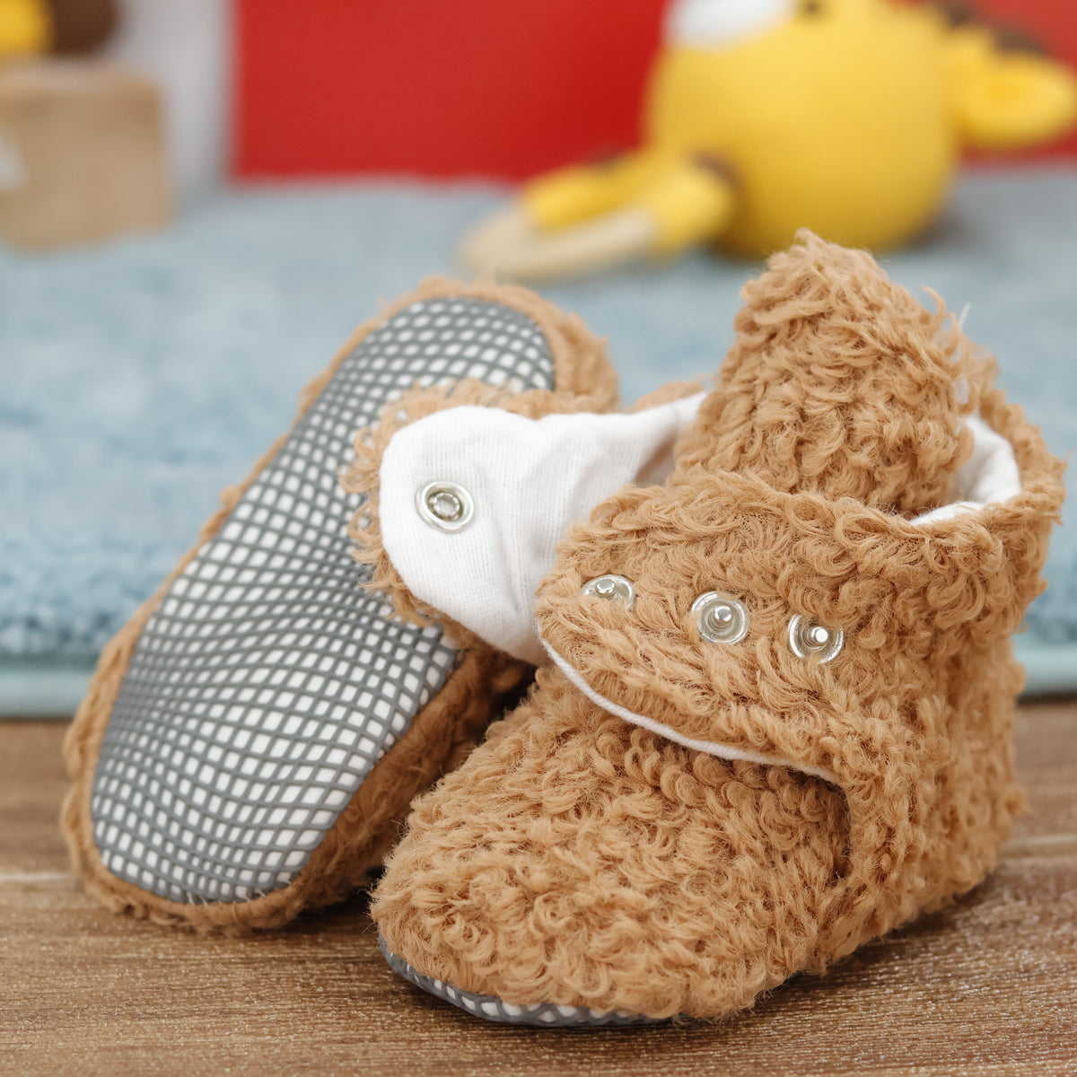 Close-up of light brown teddy-texture baby booties with snap straps and gray grip soles, showing one sole and an open snap strap.