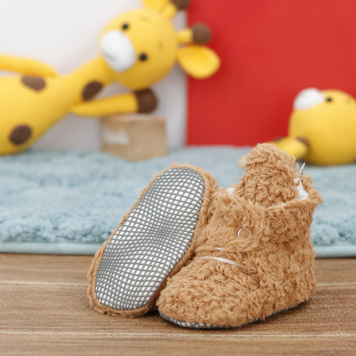 Light brown teddy-texture baby booties with snap straps and gray grip soles arranged on a surface with one sole facing the camera.