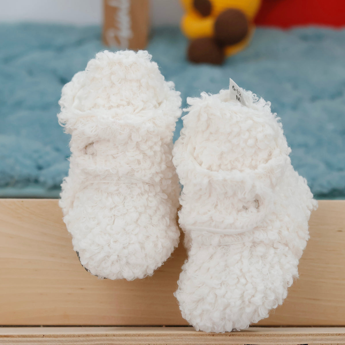 Front view of a pair of off-white teddy-texture baby booties with snap straps and gray grip soles.