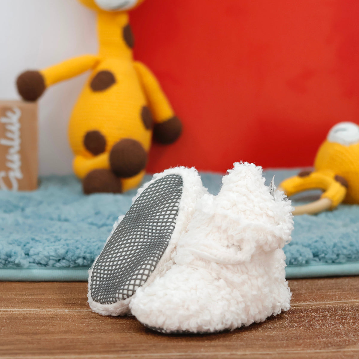 Off-white teddy-texture baby booties with snap straps and gray grip soles arranged on a surface with one sole facing the camera.