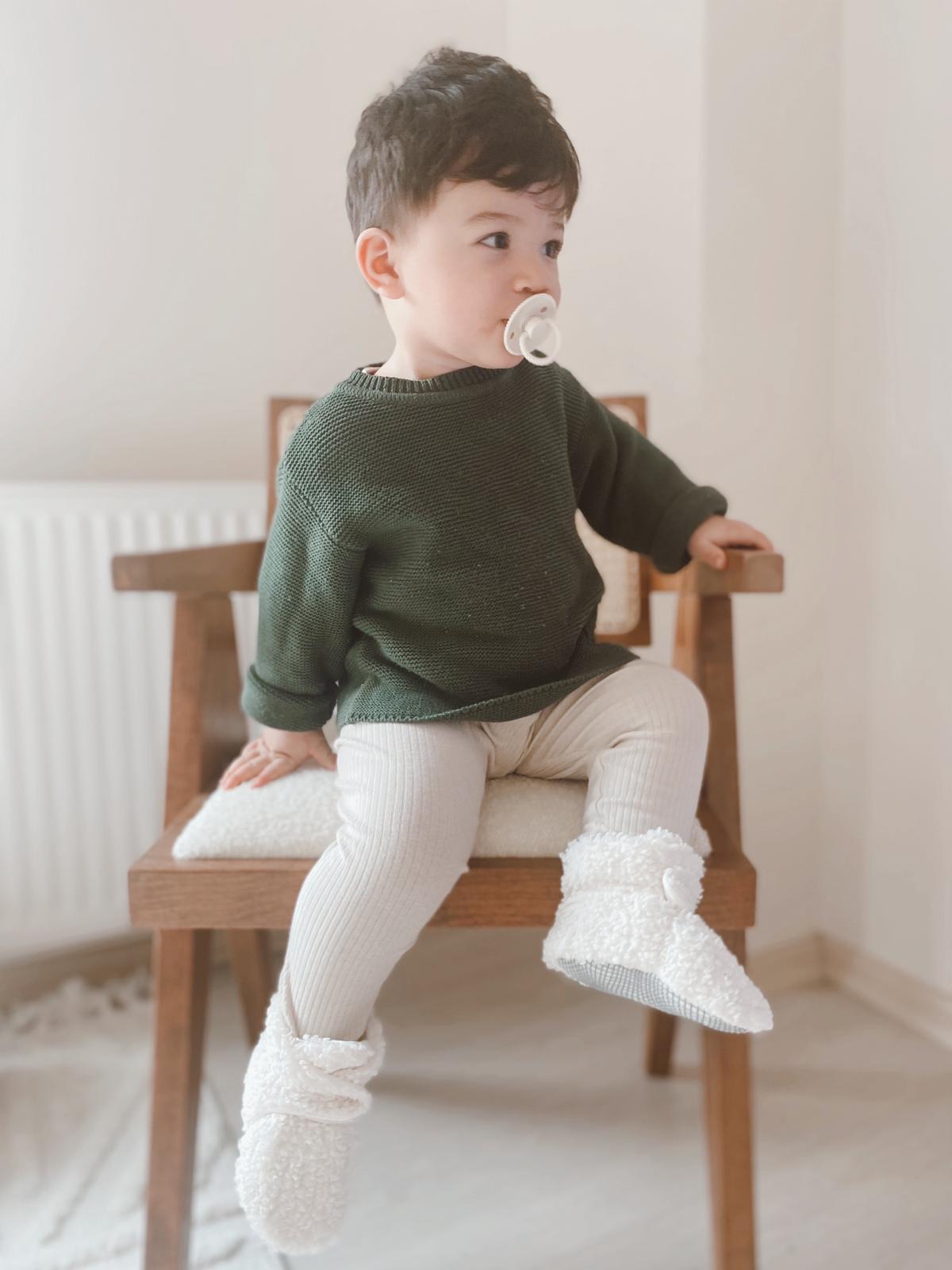 Child in a green top seated on a chair wearing white teddy booties.