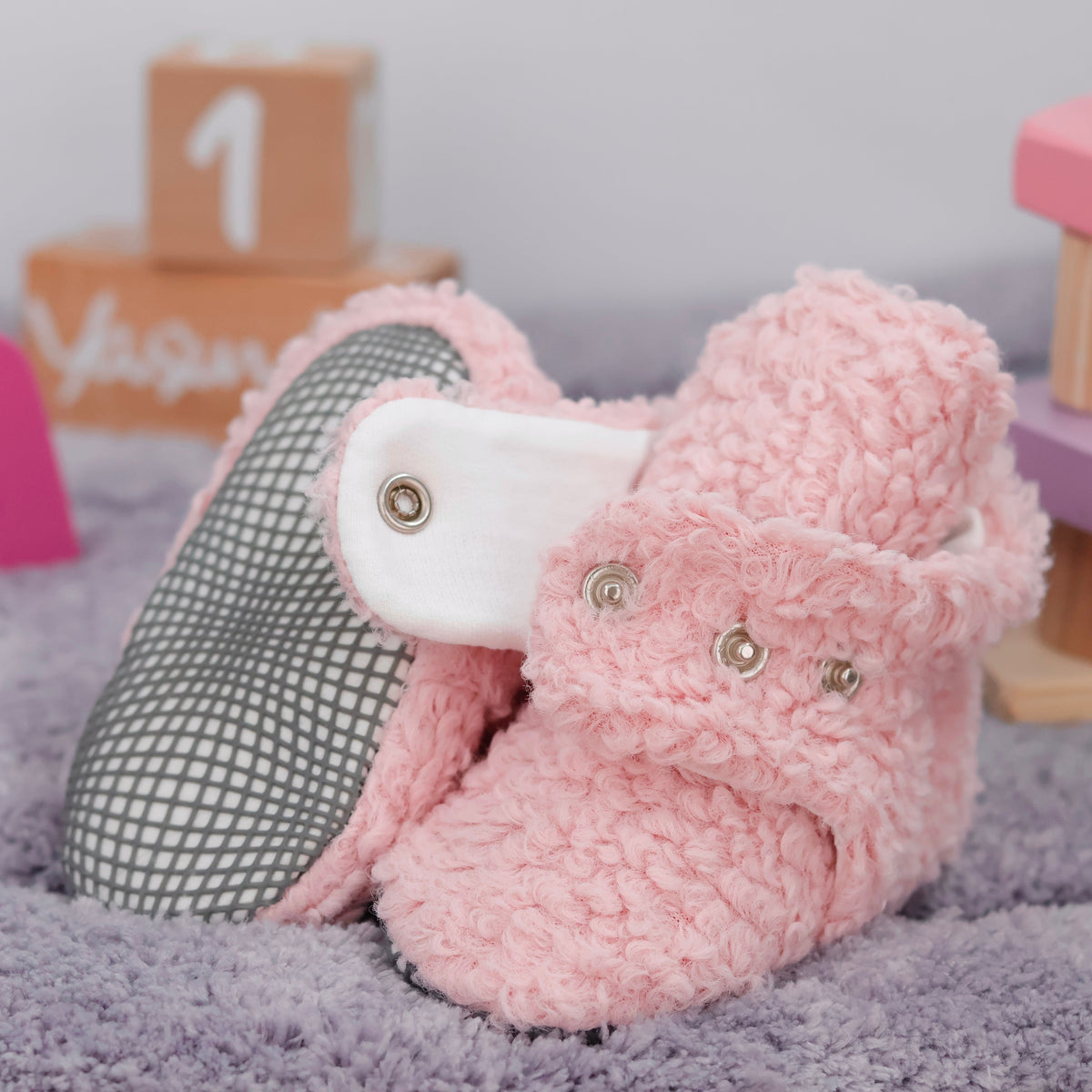 Close-up of pink teddy-texture baby booties with snap straps and gray grip soles, showing one sole and an open snap strap.