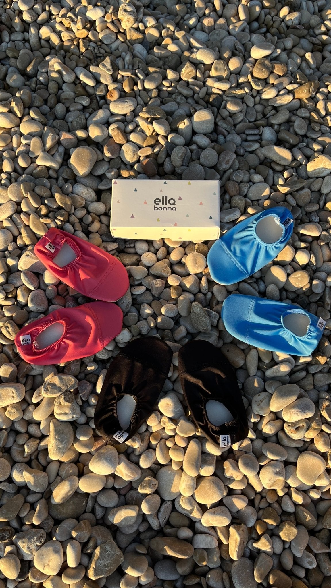 Red, black and blue water shoes arranged on a pebble beach around an Ella Bonna card.