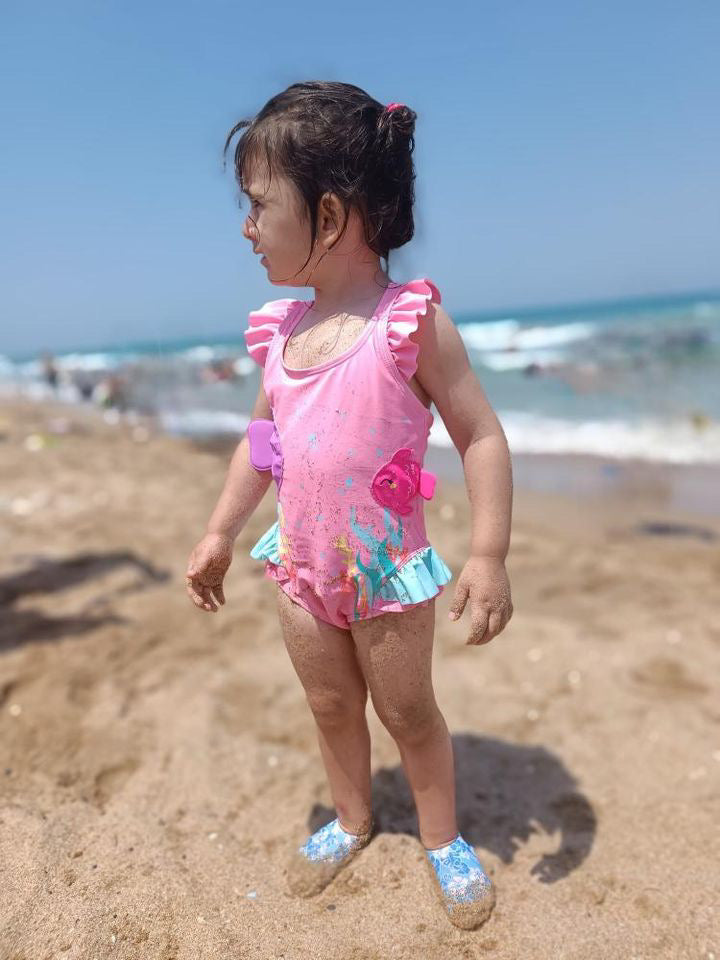 Child in a pink swimsuit standing on a beach wearing blue floral water shoes.