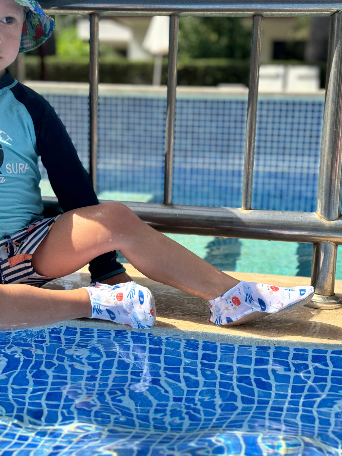 Child seated at a pool edge wearing crab-print water shoes.