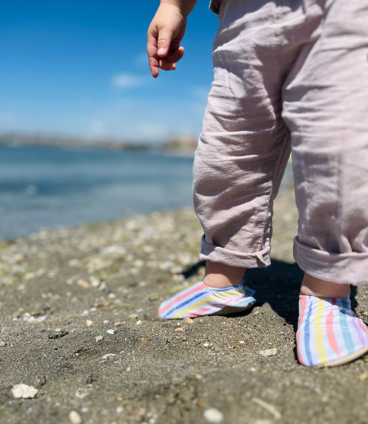 Close-up of striped water shoes worn with gray trousers on a beach.