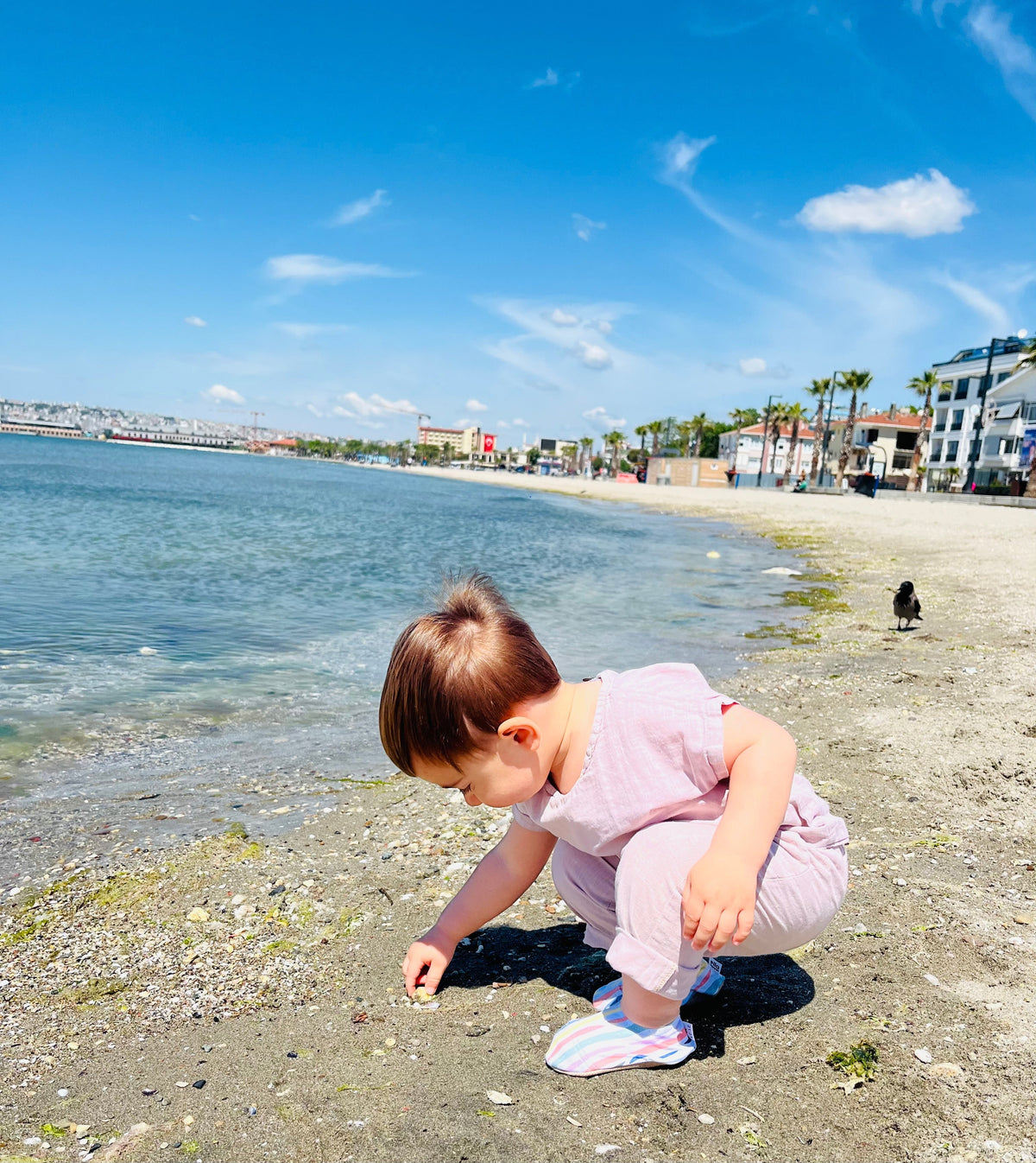 Child crouching at the shoreline wearing striped water shoes.