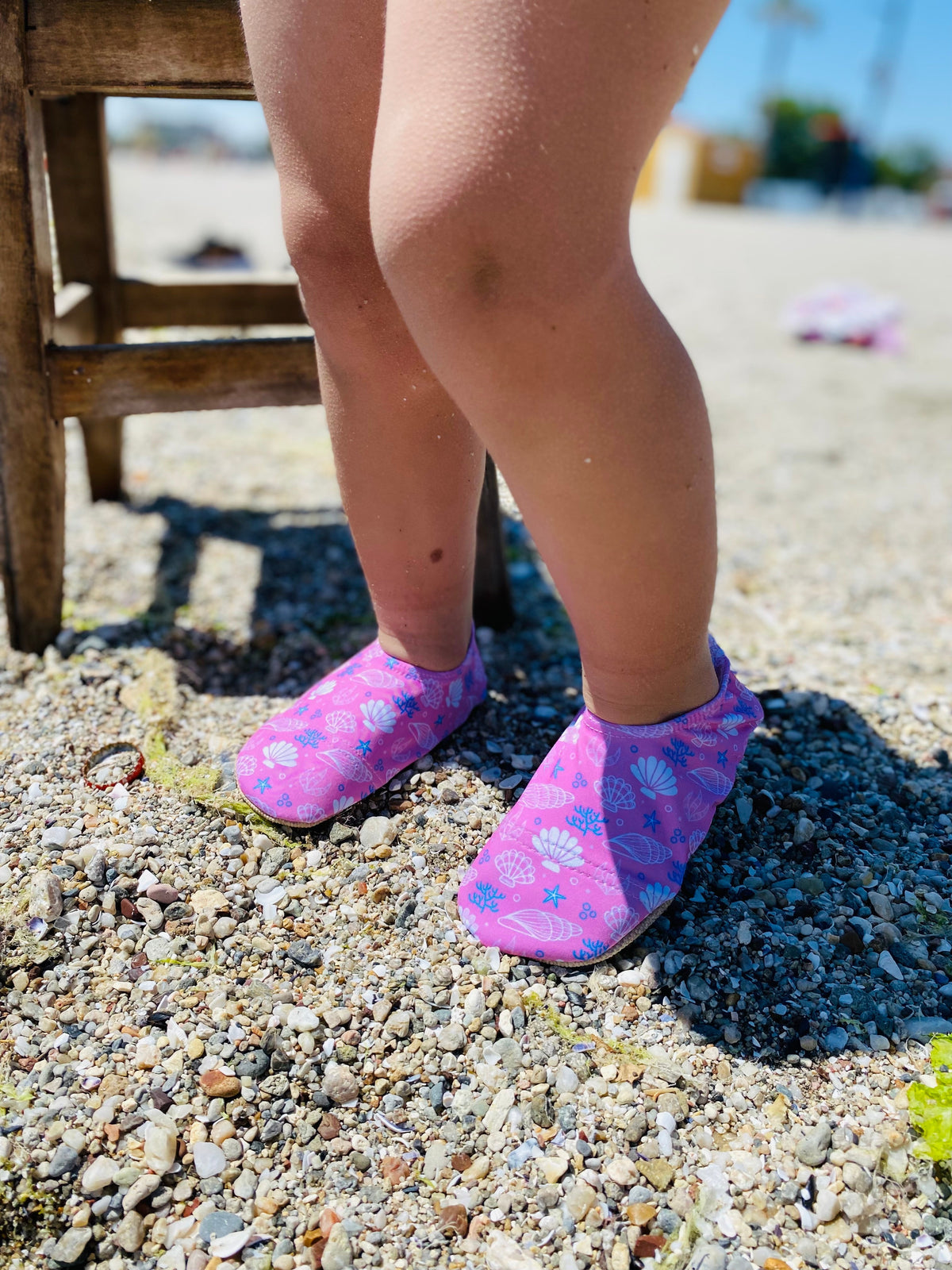 Pink shell-print water shoes worn while standing on a pebble beach.