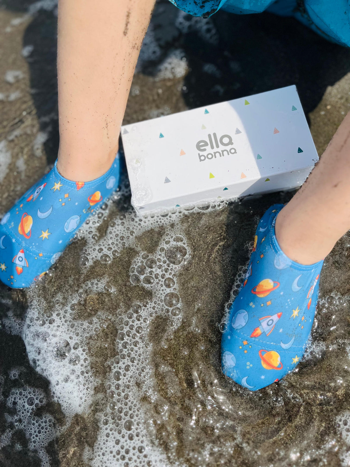 Space-print water shoes worn in shallow surf beside an Ella Bonna card.