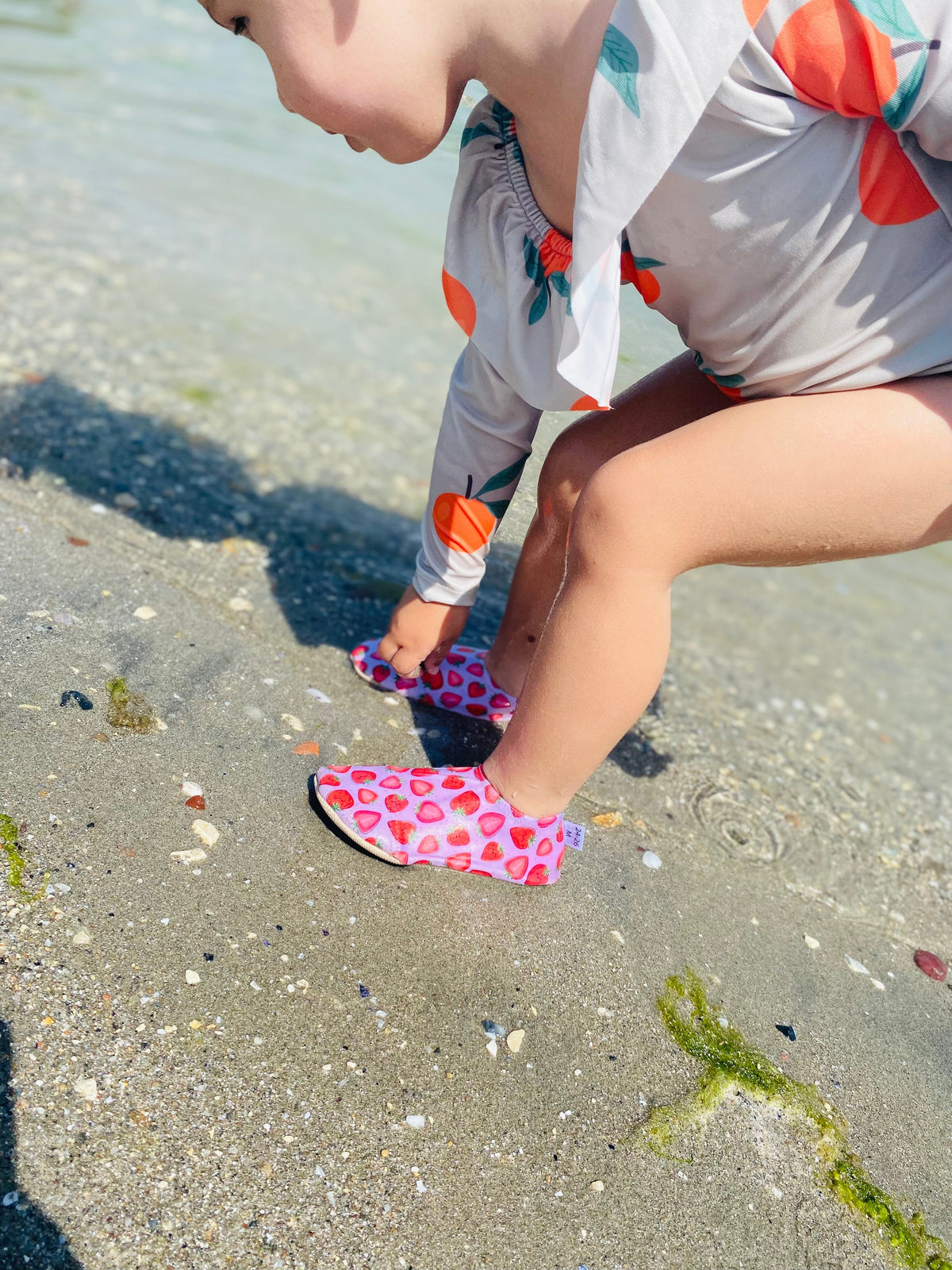 Child seated on sand wearing strawberry-print water shoes.