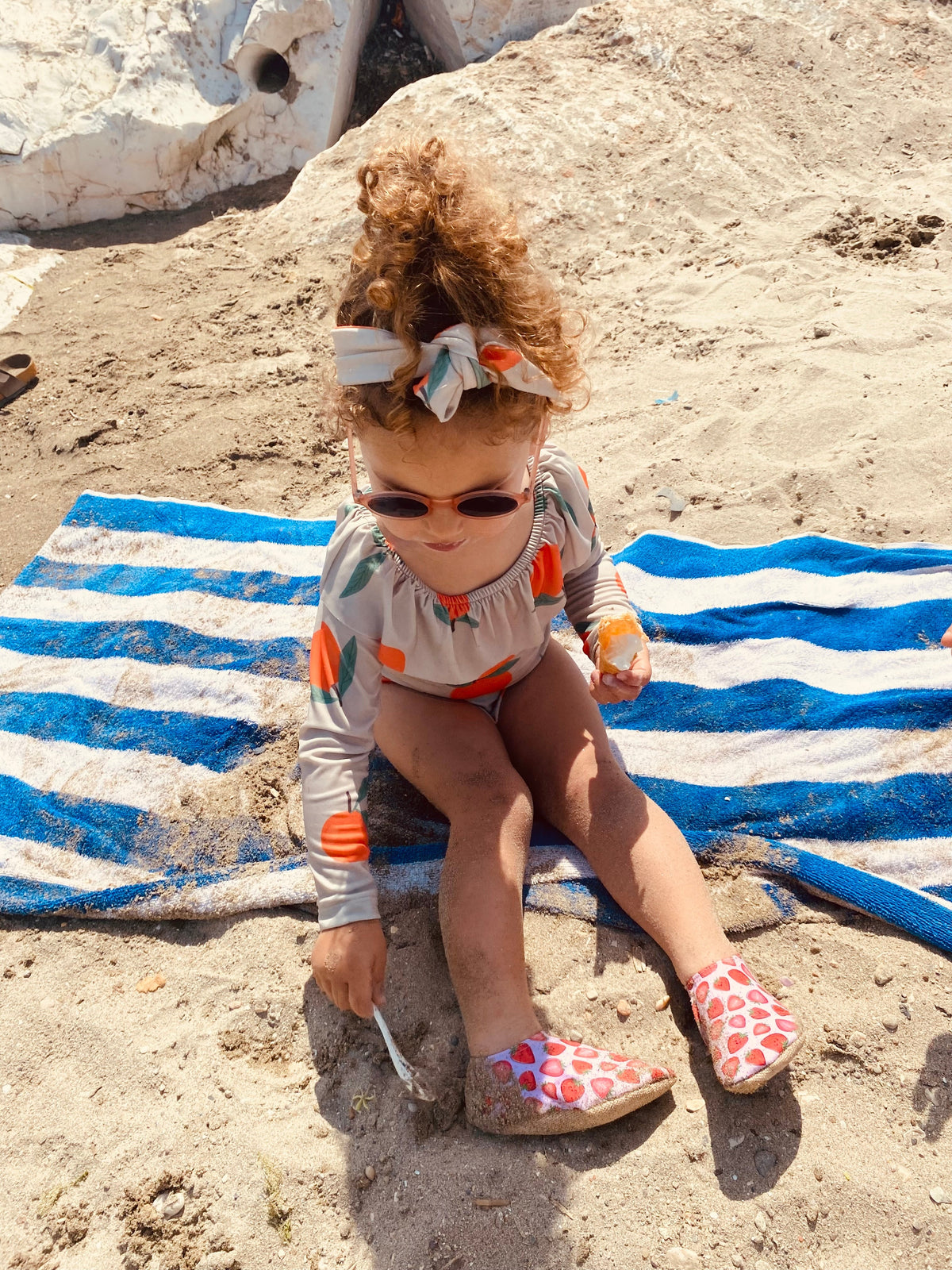 Child seated on sand wearing strawberry-print water shoes, front view.