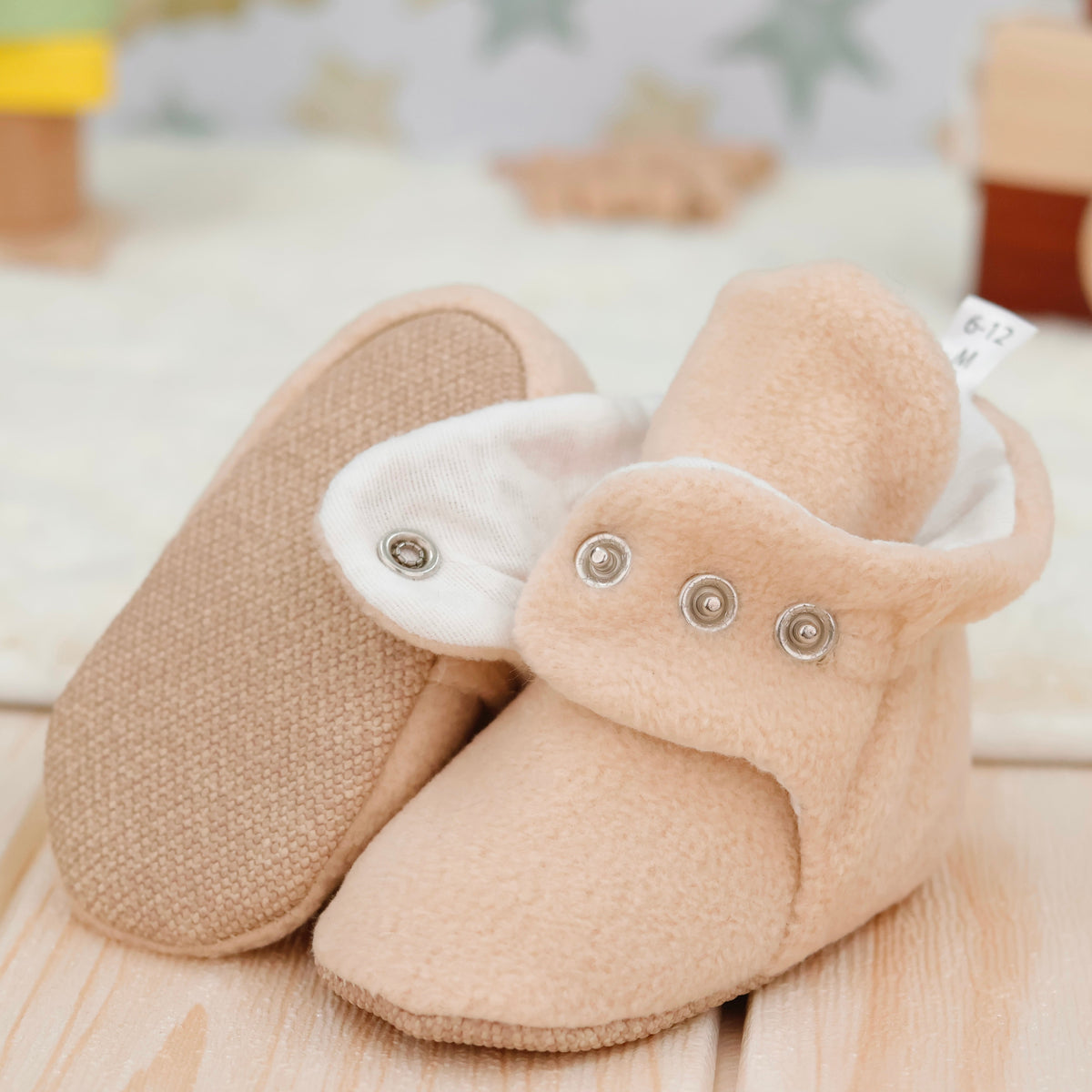 Close-up of beige fleece-texture baby booties with snap straps and tan soles, showing one sole and an open snap strap.