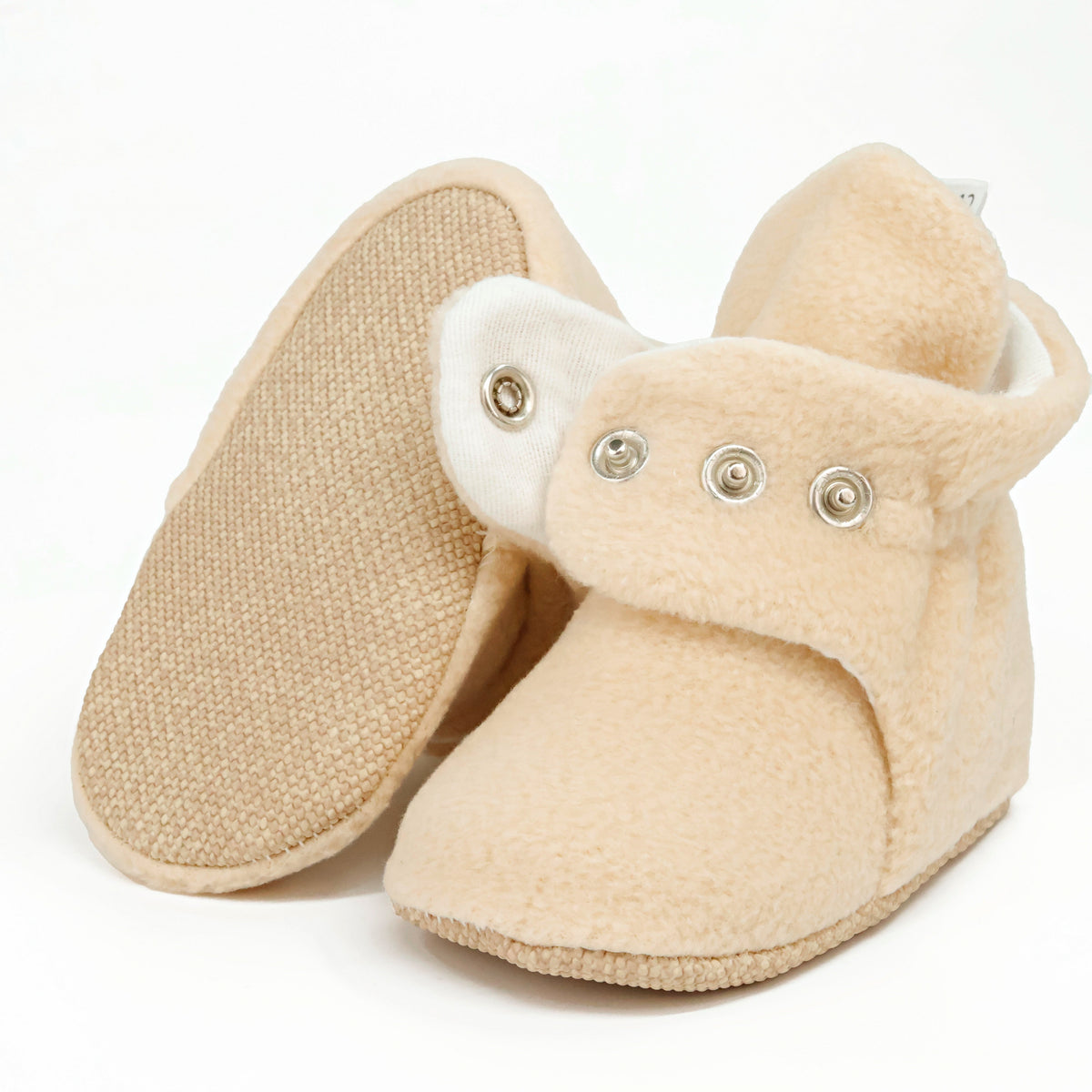 Beige fleece-texture baby booties with snap straps and tan soles, showing one sole and an open snap strap on white.