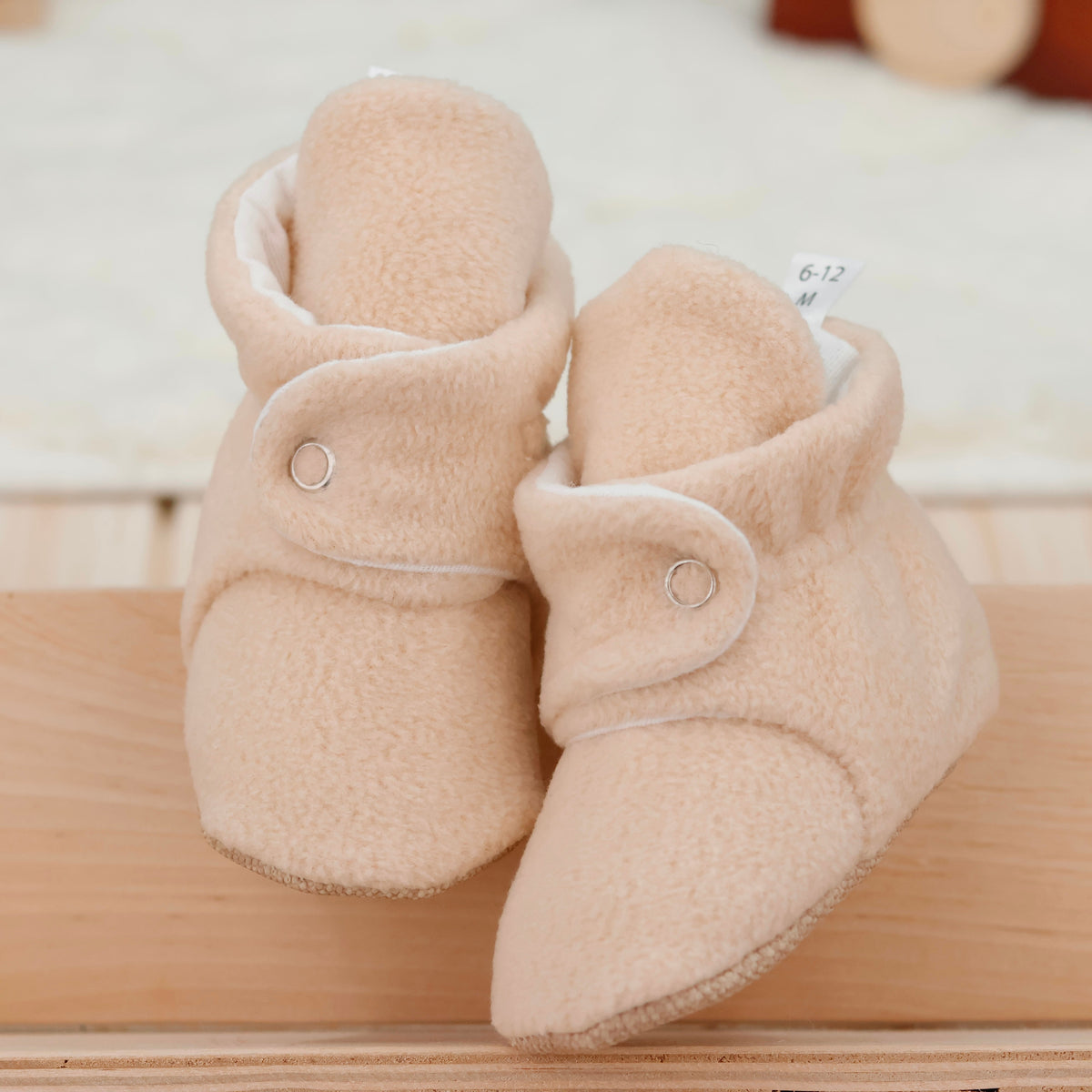 Front view of a pair of beige fleece-texture baby booties with snap straps and tan soles.