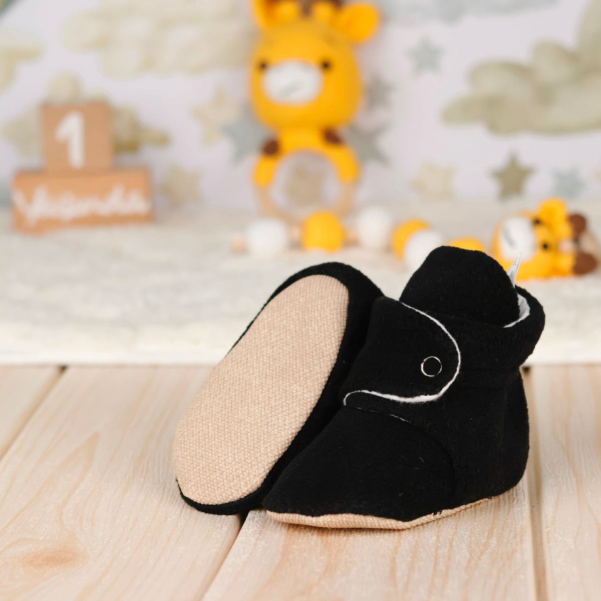 Black fleece-texture baby booties with snap straps and tan soles arranged on a surface with one sole facing the camera.