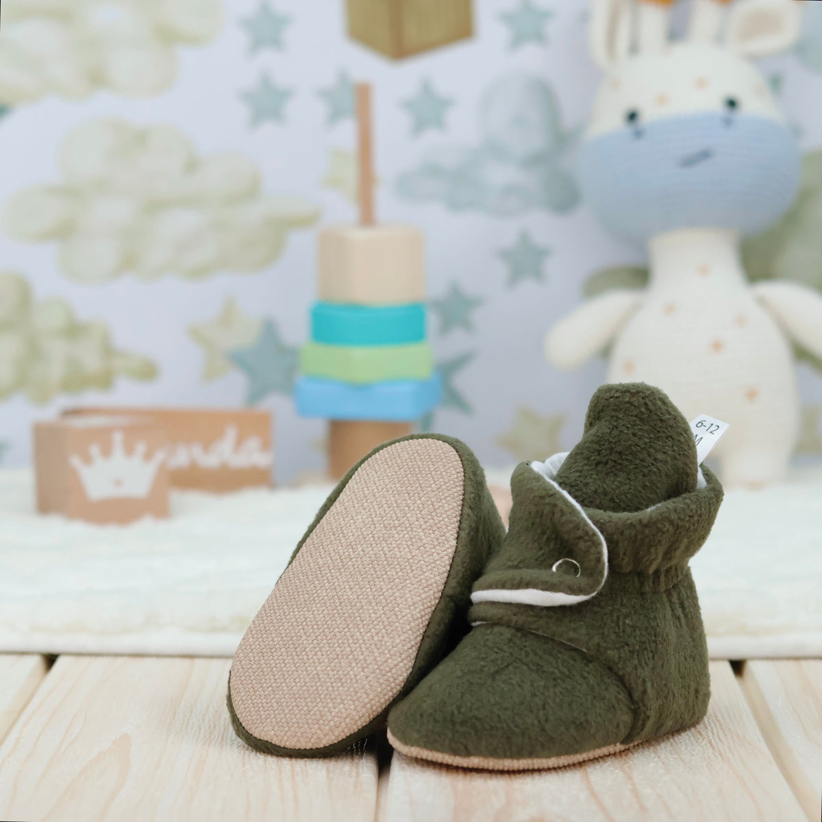 Olive green fleece-texture baby booties with snap straps and tan soles arranged on a surface with one sole facing the camera.
