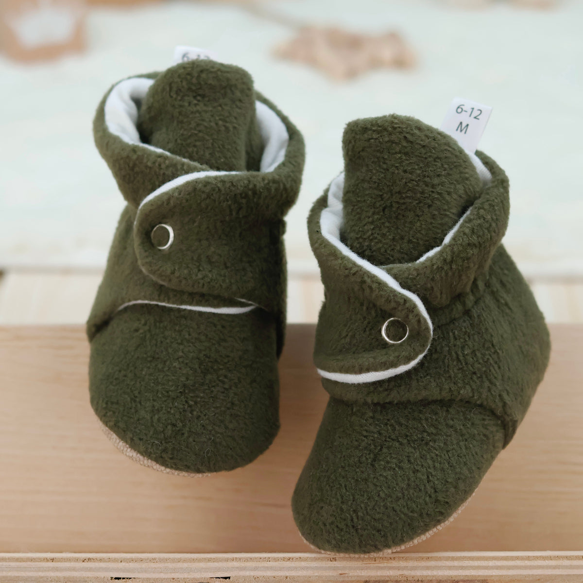 Front view of a pair of olive green fleece-texture baby booties with snap straps and tan soles.
