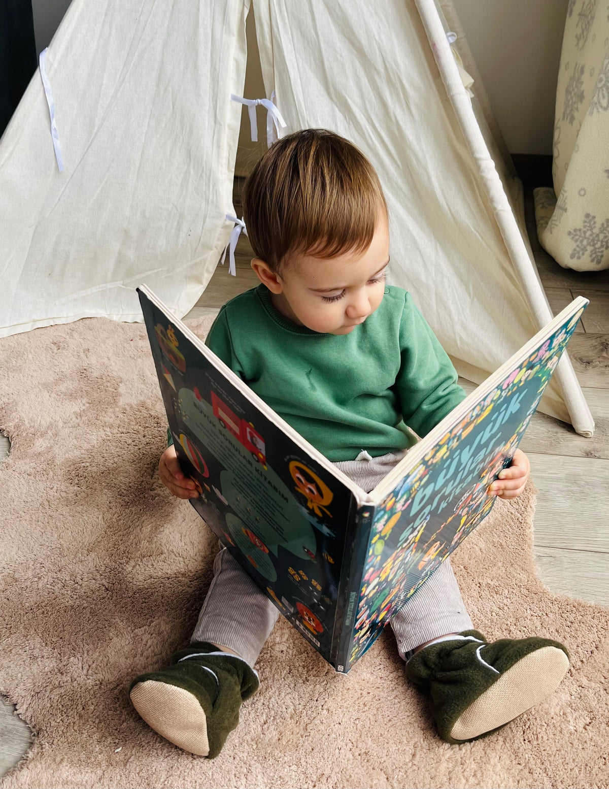 Child seated reading a book while wearing olive booties.