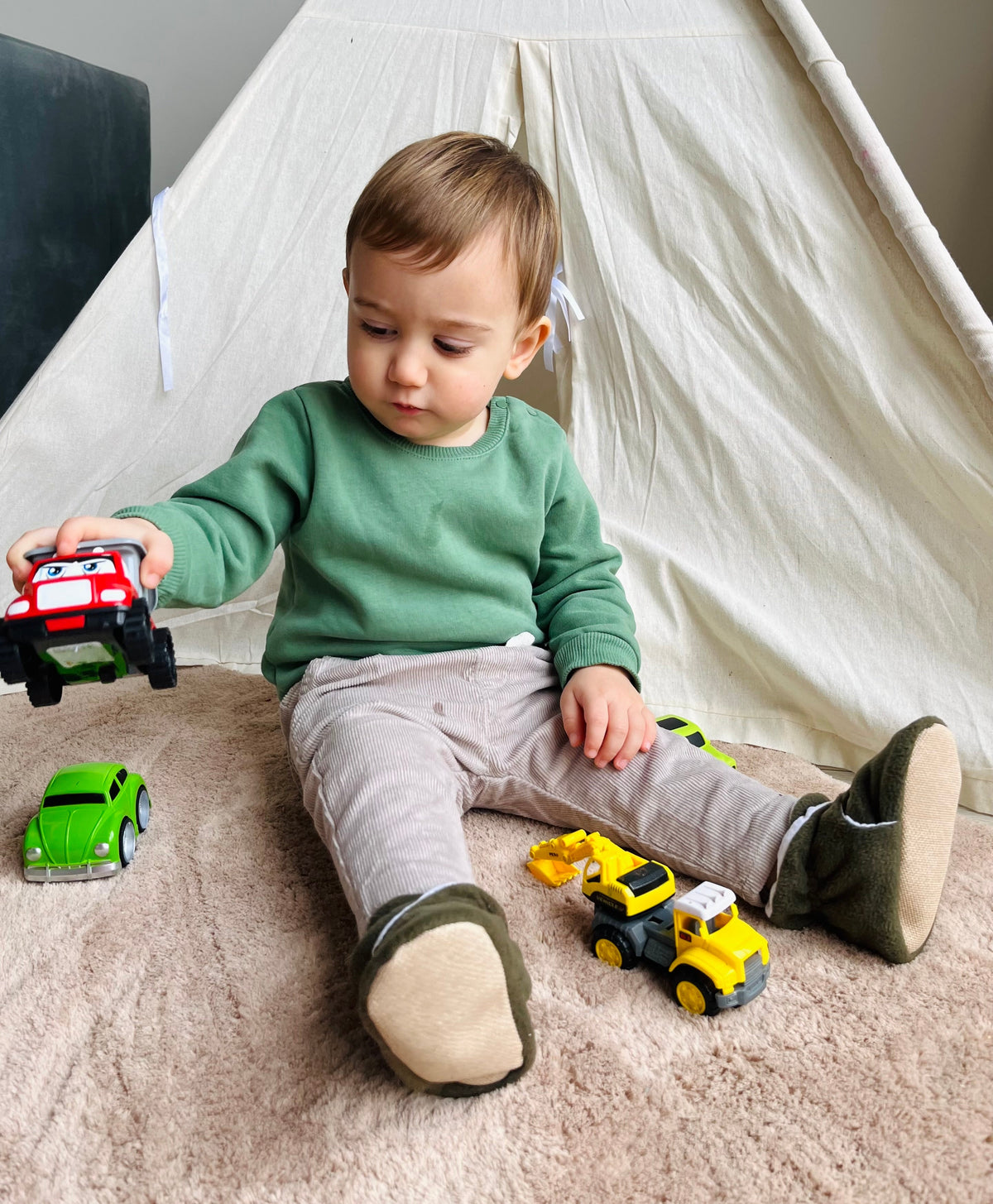 Child seated with toy vehicles wearing olive booties.