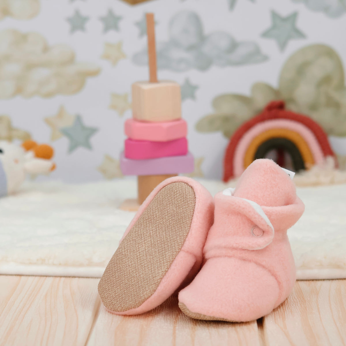 Pale pink fleece-texture baby booties with snap straps and tan soles arranged on a surface with one sole facing the camera.