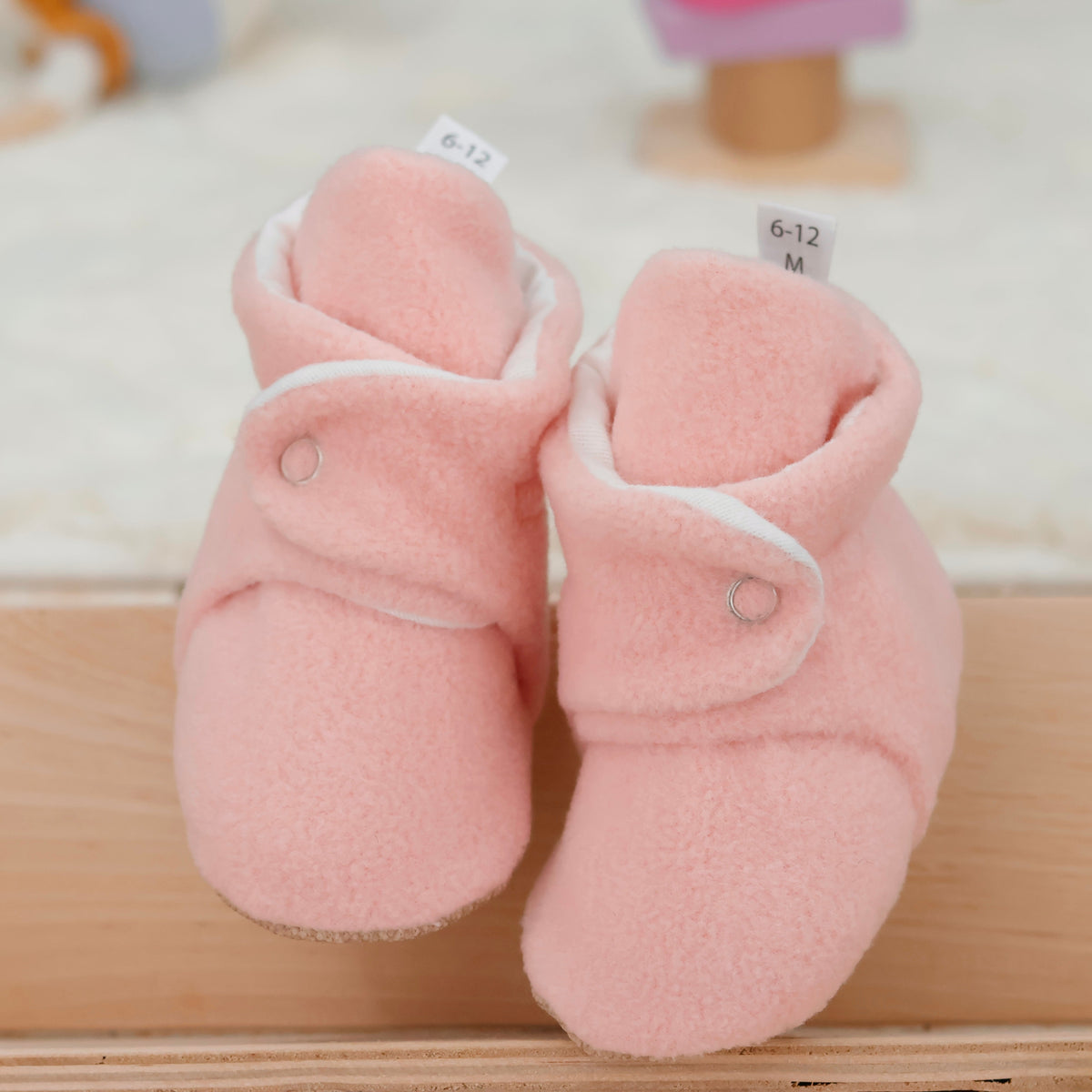 Front view of a pair of pale pink fleece-texture baby booties with snap straps and tan soles.