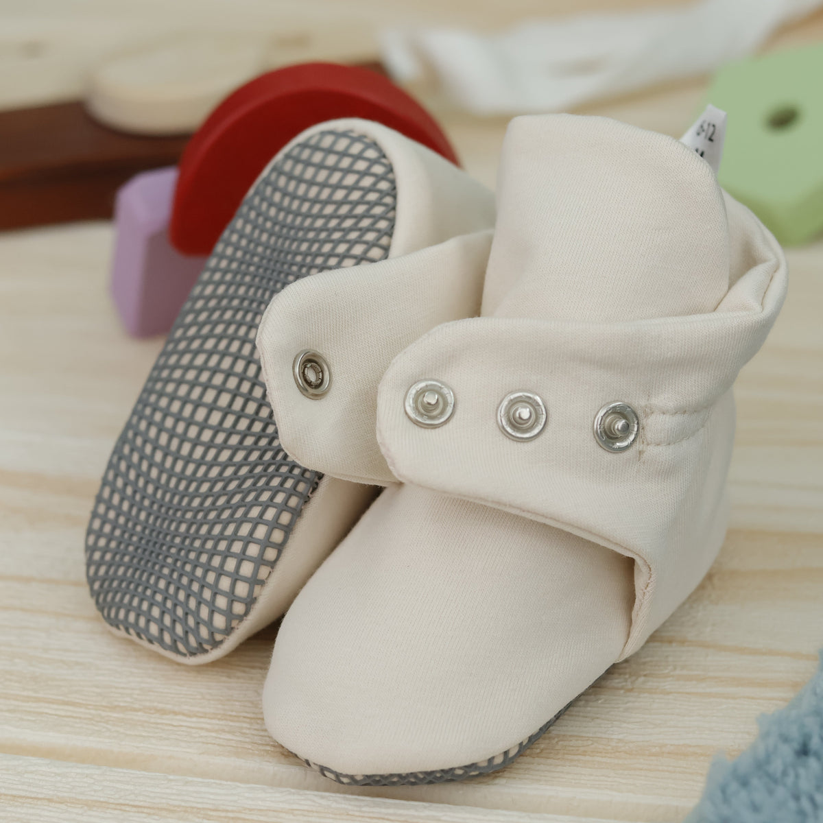 Close-up of cream fabric baby booties with snap straps and gray grip soles, showing one sole and an open snap strap.