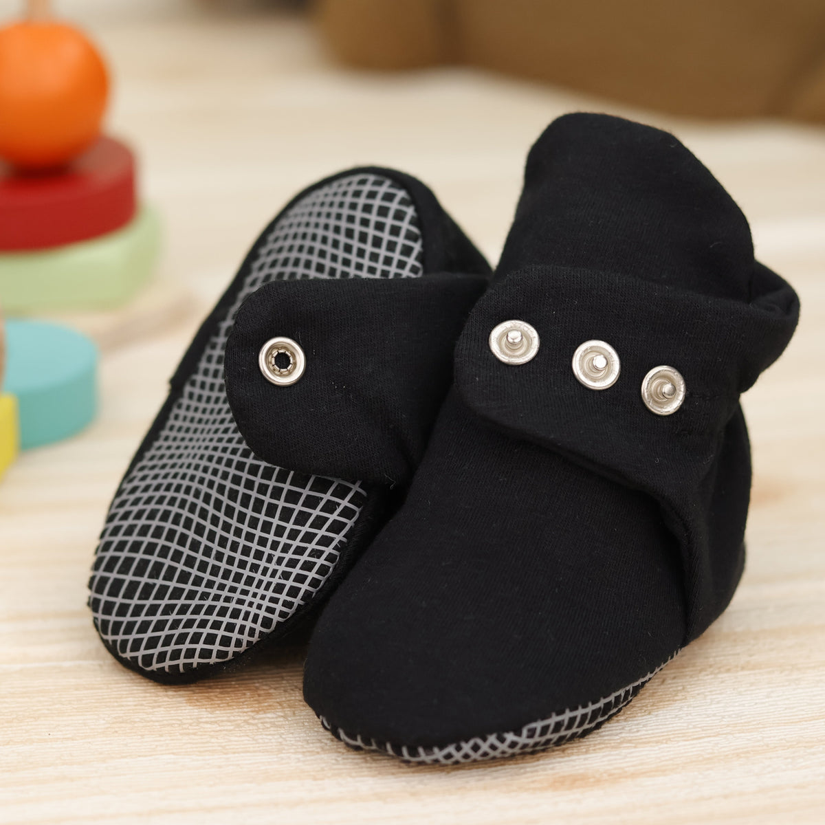 Close-up of black fabric baby booties with snap straps and gray grip soles, showing one sole and an open snap strap.