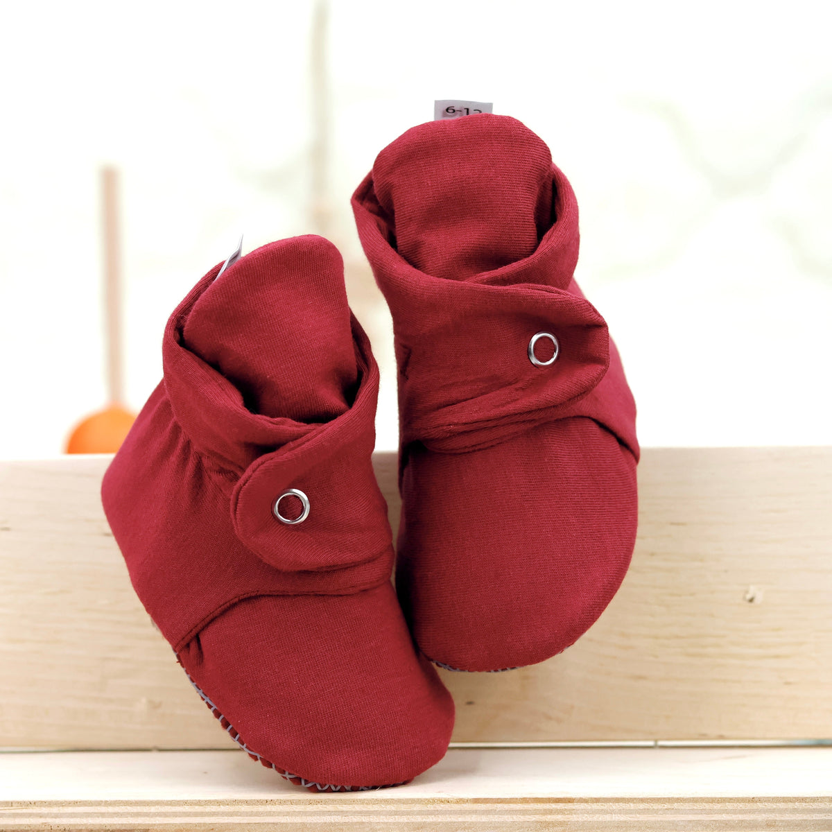 Front view of a pair of burgundy fabric baby booties with snap straps and gray grip soles.