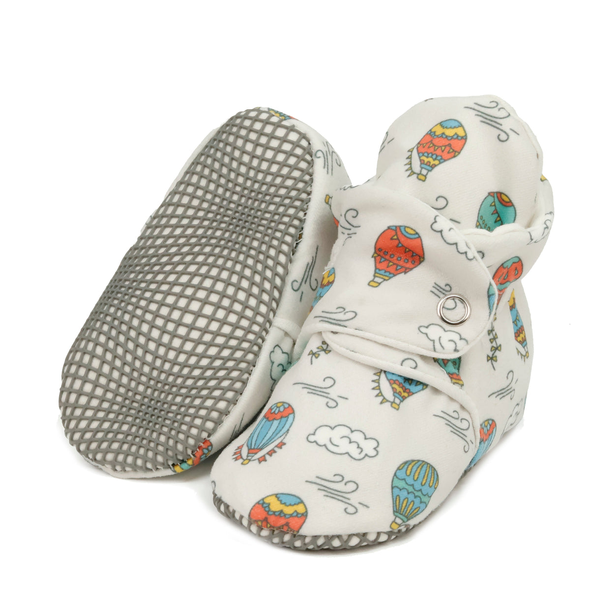 Cream baby booties with colorful hot-air balloons, clouds and snap straps, with one sole facing the camera on a white background.