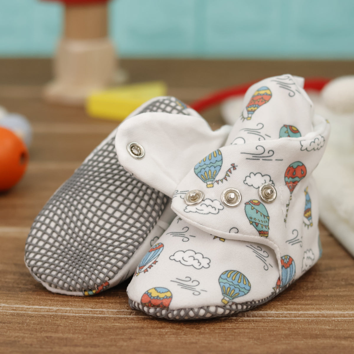 Close-up of cream baby booties with colorful hot-air balloons, clouds and snap straps, showing one sole and an open snap strap.