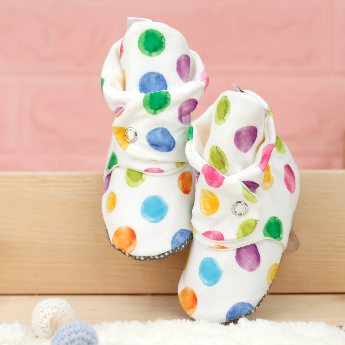 Front view of a pair of white baby booties with multicolored watercolor dots and snap straps.