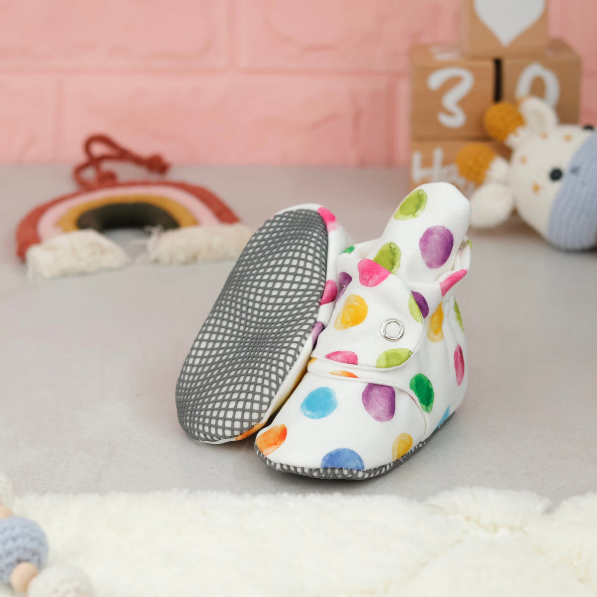 White baby booties with multicolored watercolor dots and snap straps arranged on a surface with one sole facing the camera.