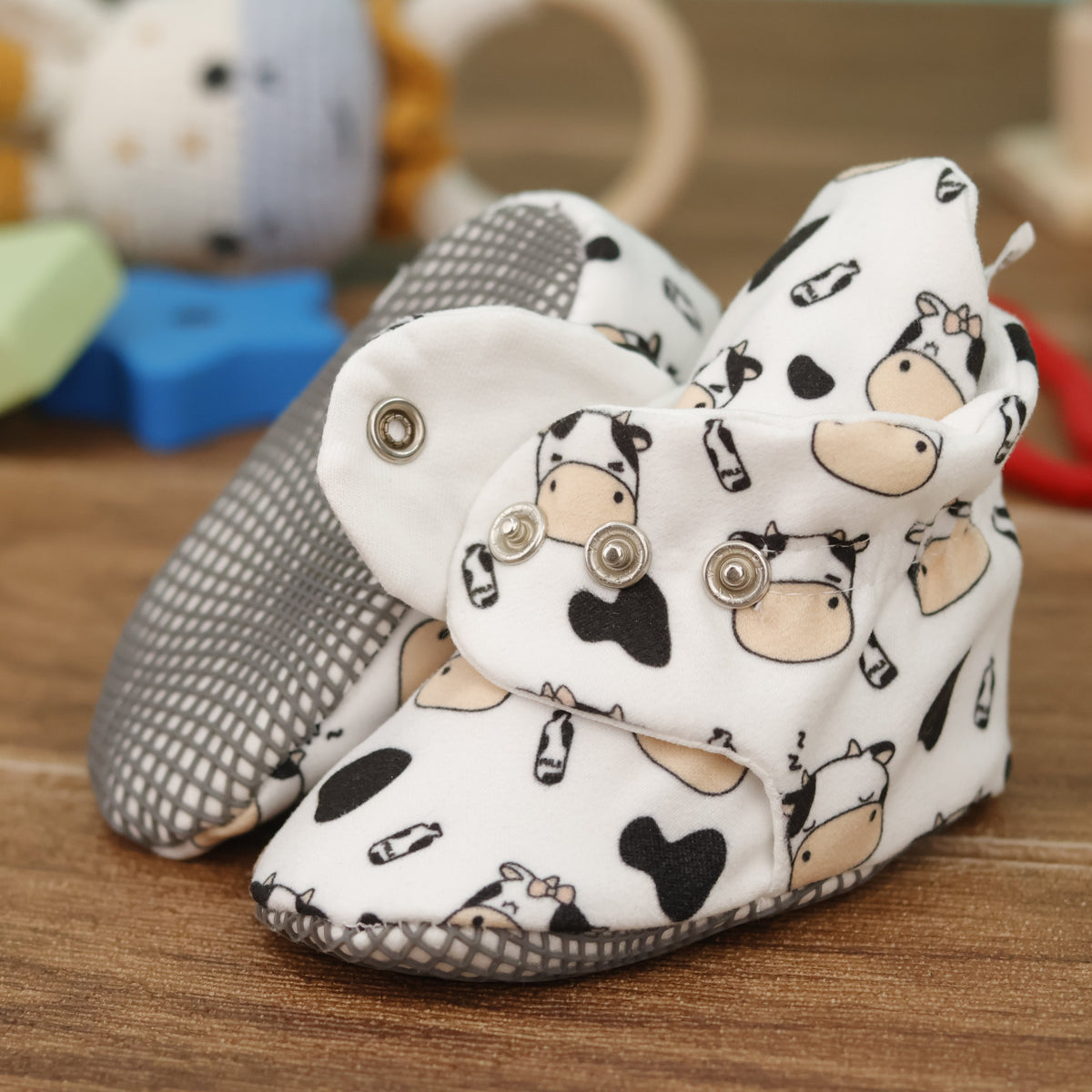 Close-up of cream baby booties with cartoon cows, black patches and snap straps, showing one sole and an open snap strap.