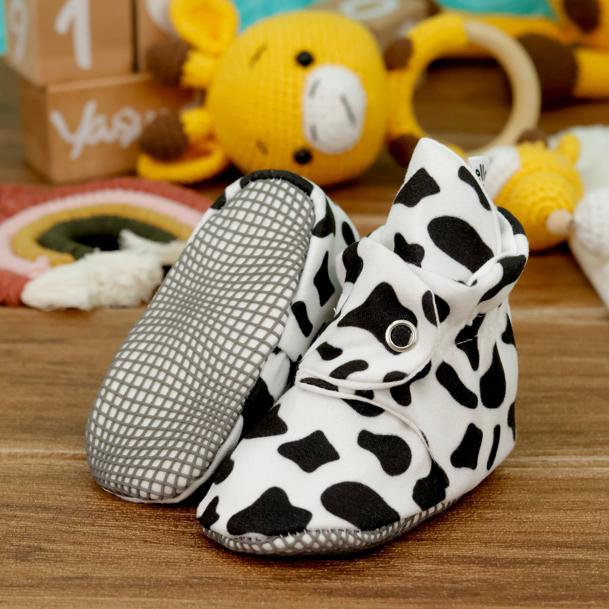 White baby booties with black cow spots and snap straps arranged on a surface with one sole facing the camera.