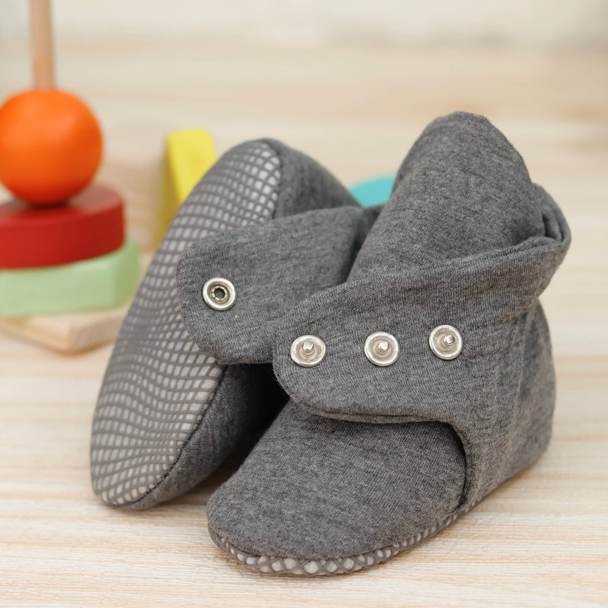 Close-up of dark gray fabric baby booties with snap straps and gray grip soles, showing one sole and an open snap strap.