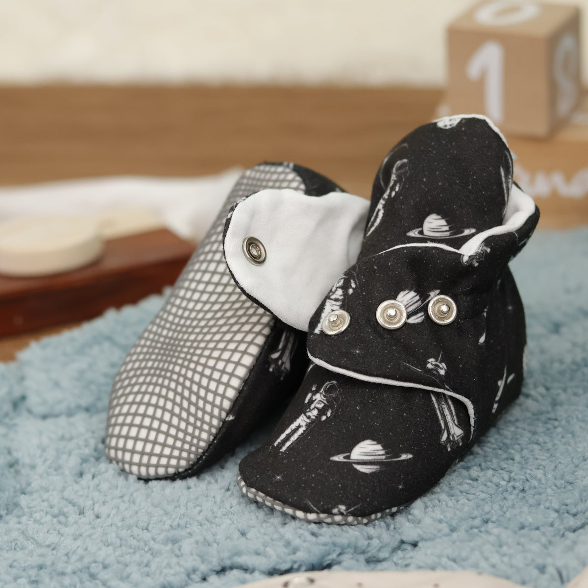 Close-up of black baby booties with white space motifs and snap straps, showing one sole and an open snap strap.