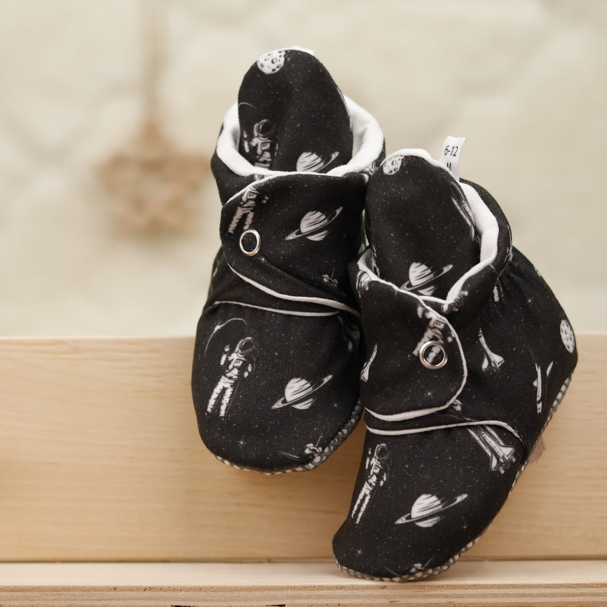 Front view of a pair of black baby booties with white space motifs and snap straps.