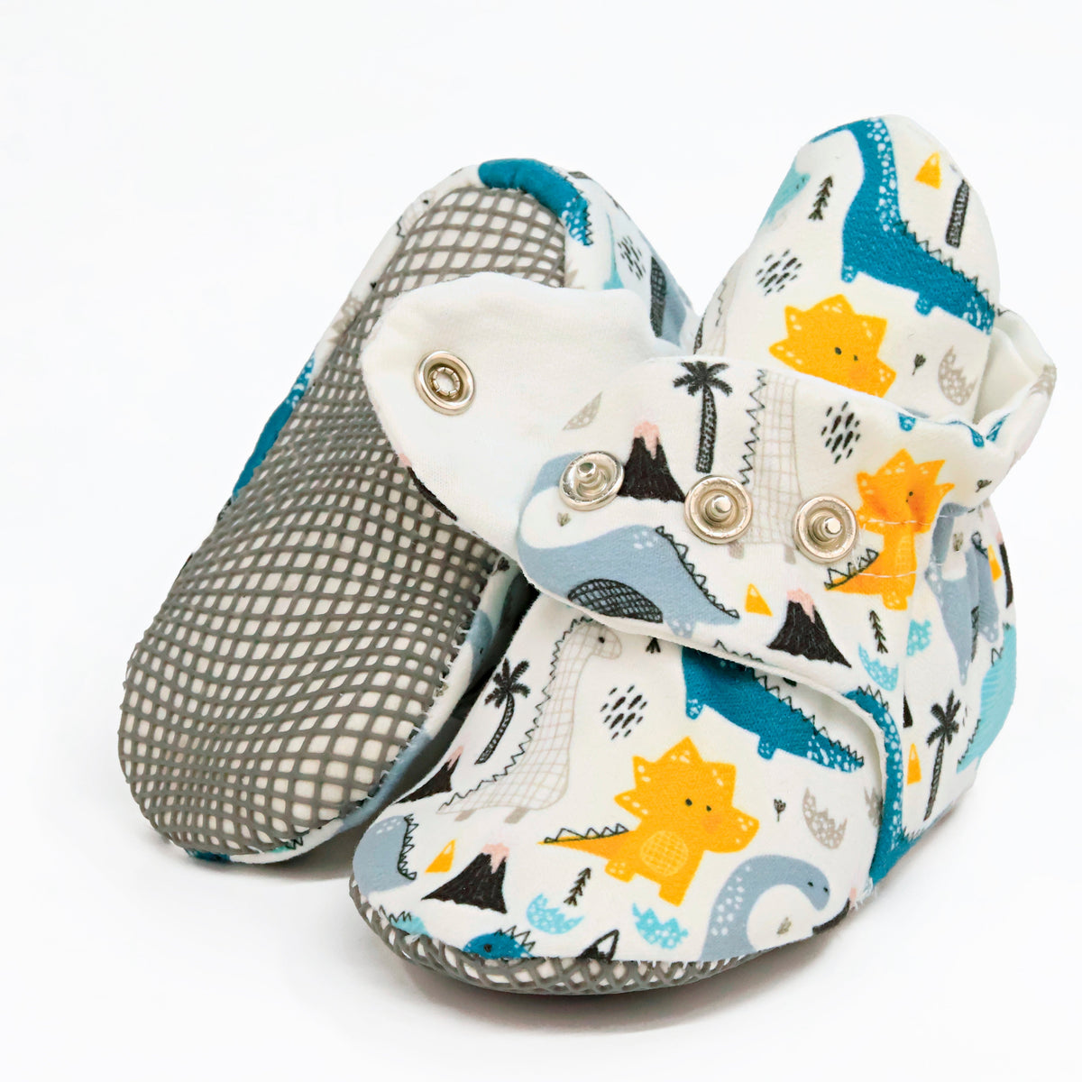 Cream baby booties with blue, gray and yellow dinosaurs and snap straps, showing one sole and an open snap strap on white.