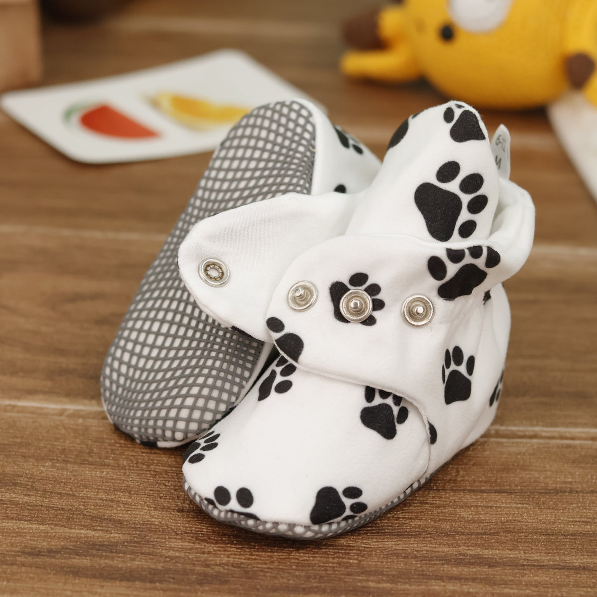 Close-up of white baby booties with black paw prints and snap straps, showing one sole and an open snap strap.