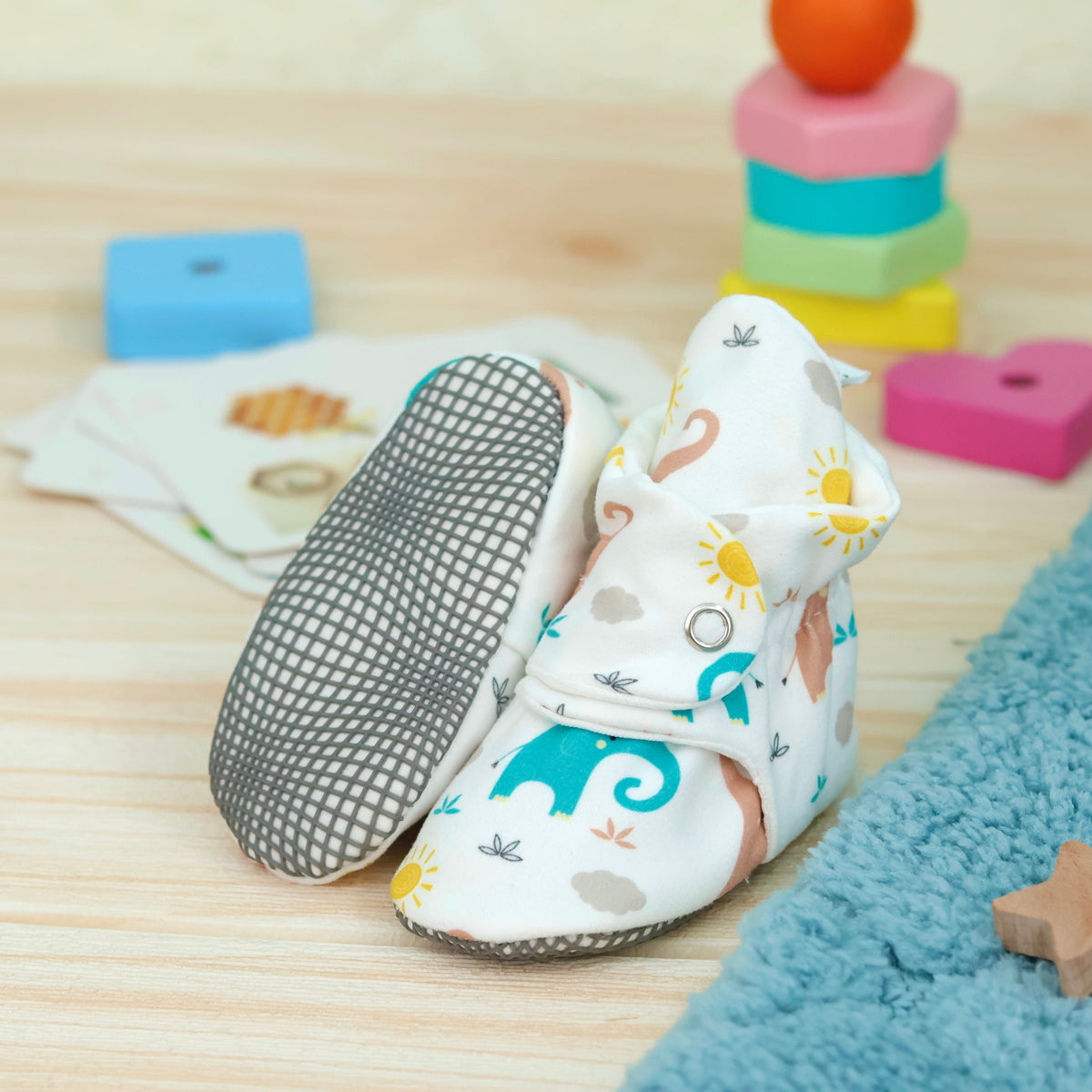 Cream baby booties with pastel elephant print and snap straps arranged on a surface with one sole facing the camera.