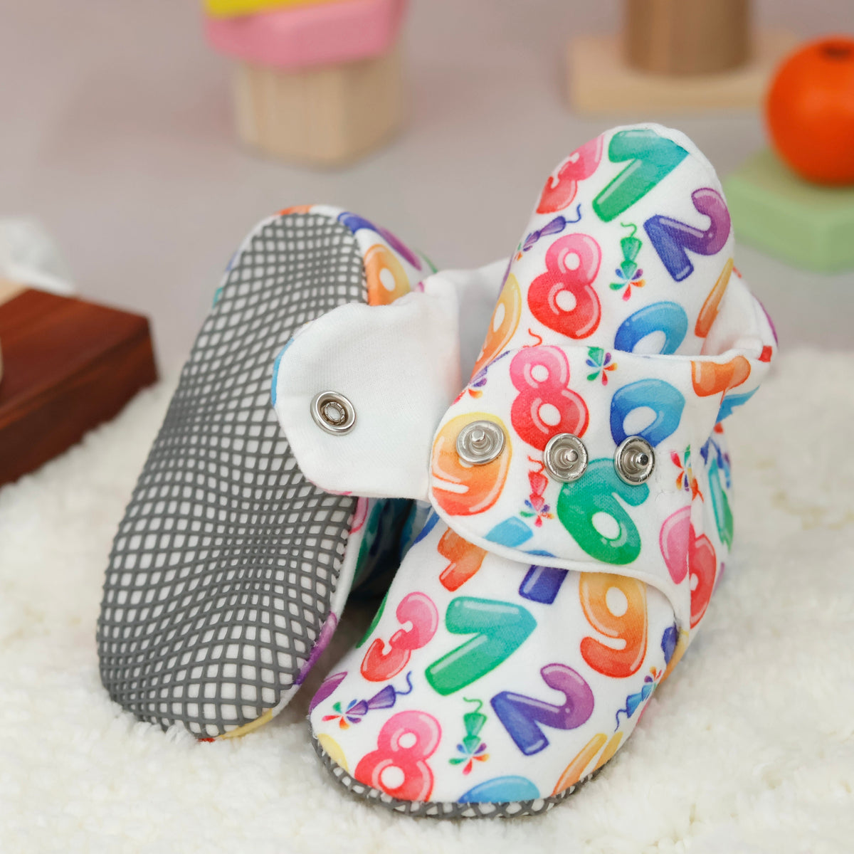 Close-up of white baby booties with colorful numbers and snap straps, showing one sole and an open snap strap.
