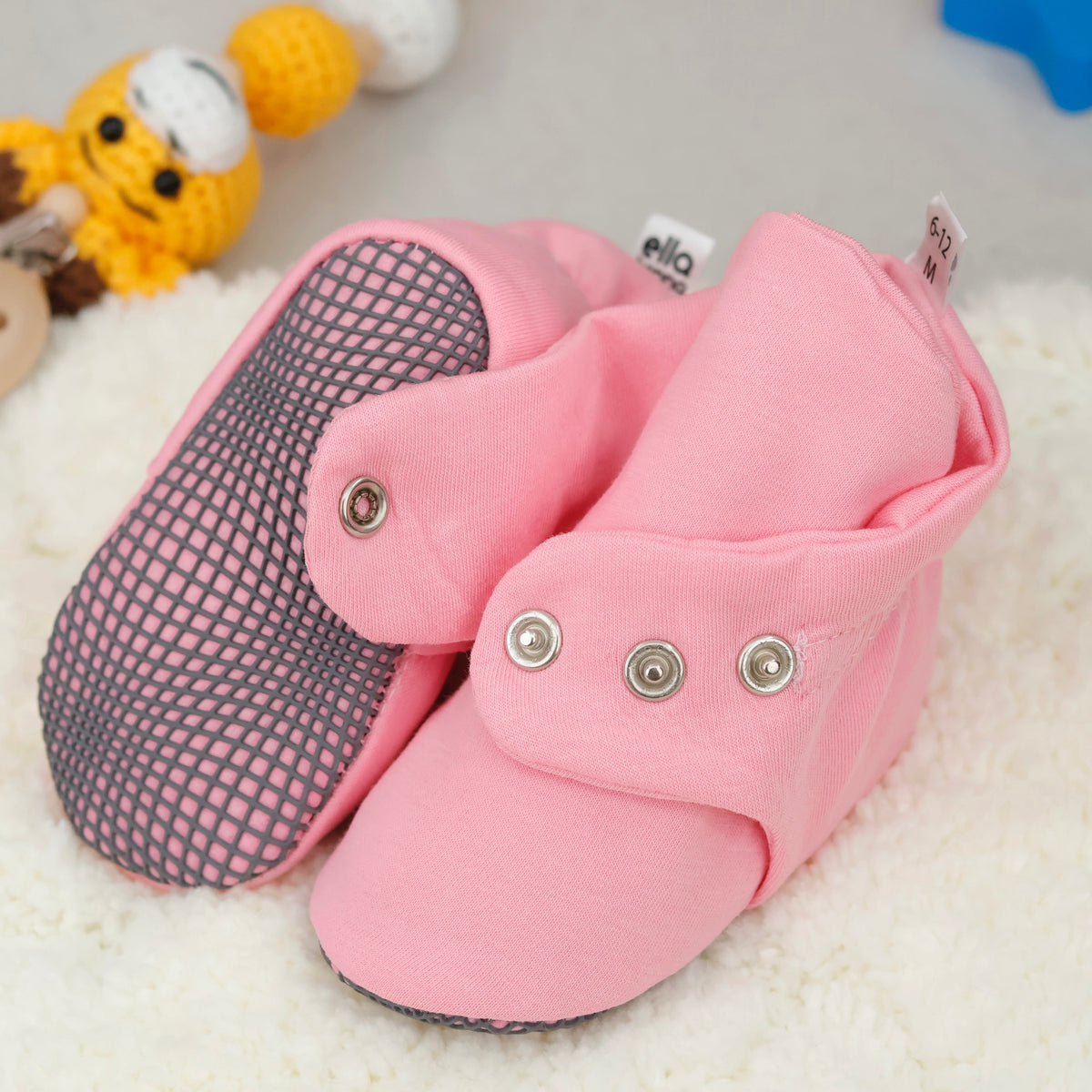 Close-up of pink fabric baby booties with snap straps and gray grip soles, showing one sole and an open snap strap.