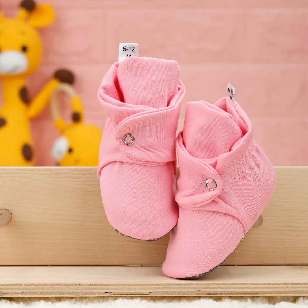 Front view of a pair of pink fabric baby booties with snap straps and gray grip soles.